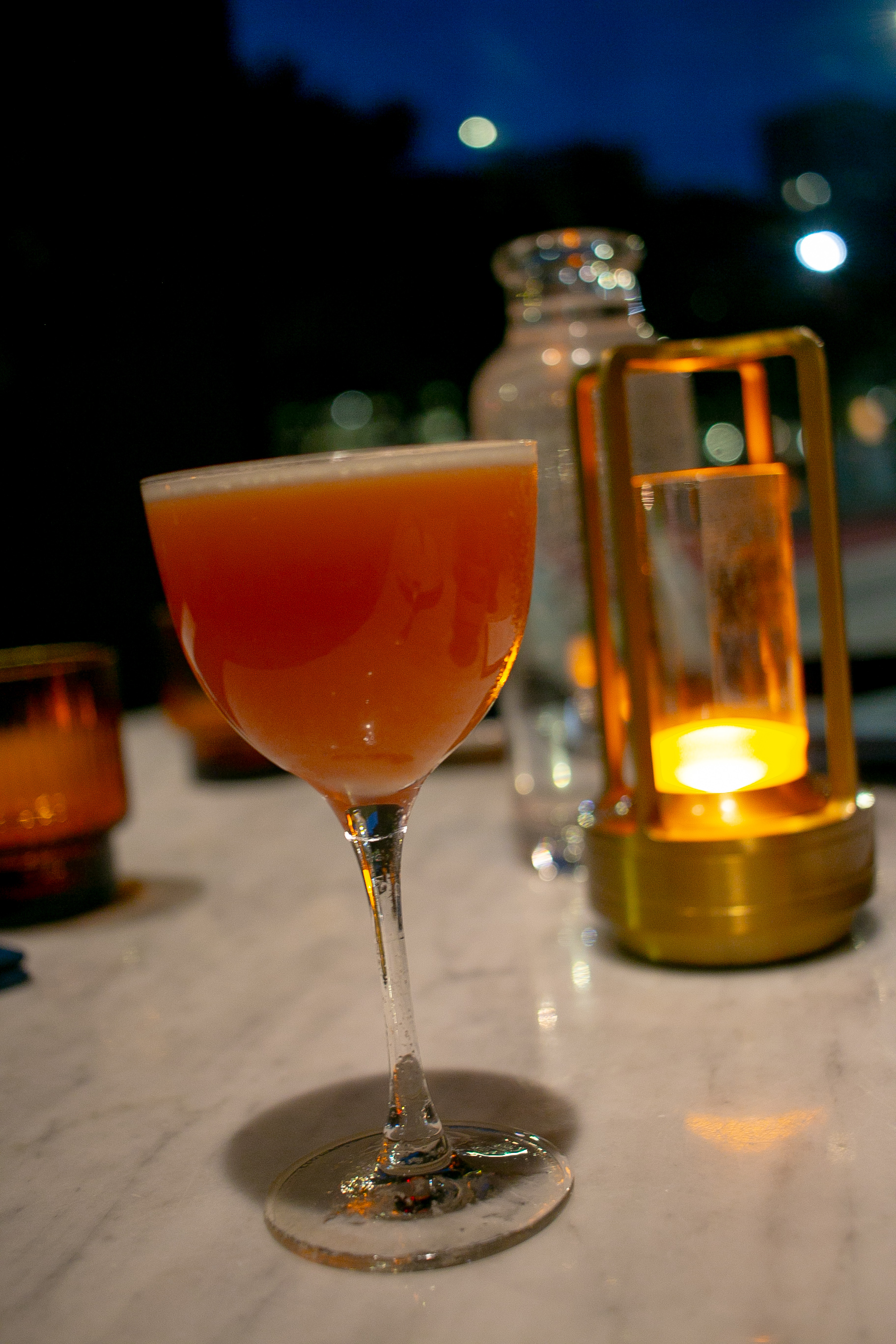 The Ticket to Paradise cocktail in a coupe glass beside a lantern at AMA