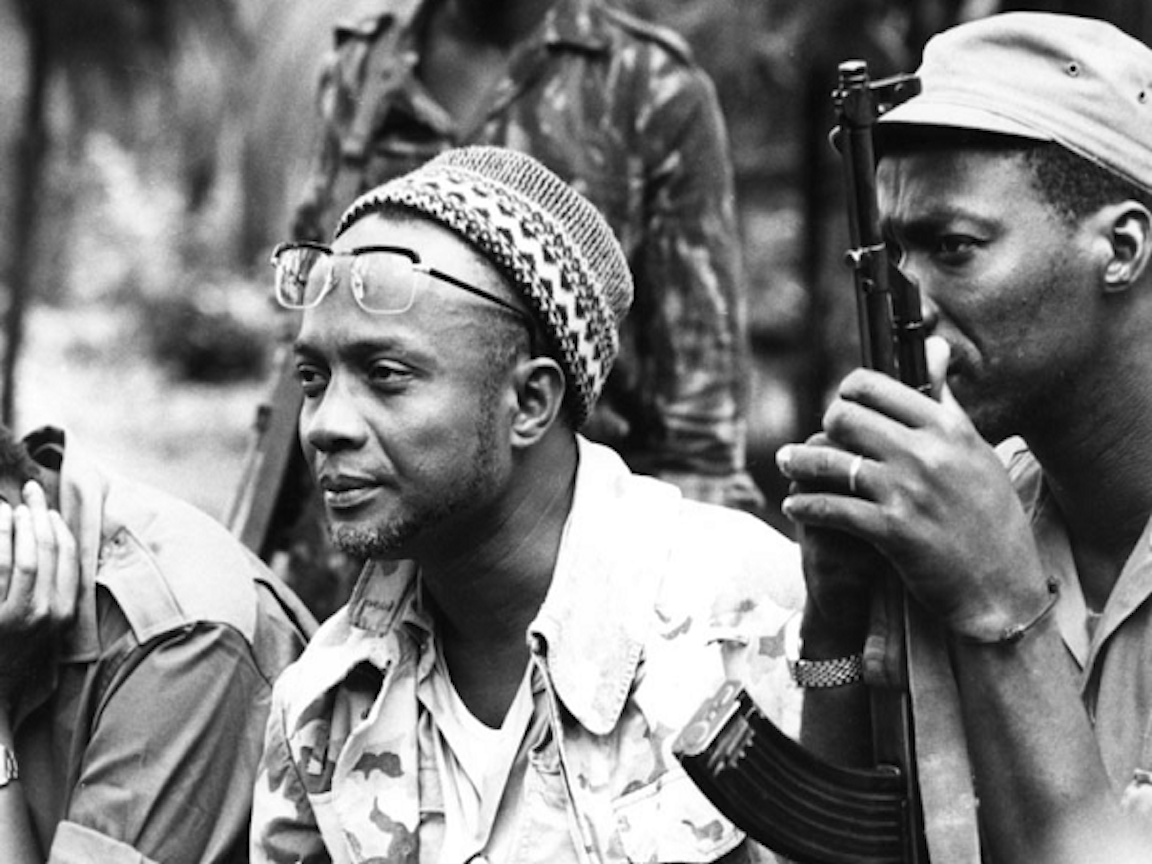 Black and white photograph of Amílcar Cabral, Cape Verdean revolutionary leader, wearing glasses and a traditional cap, with a soldier visible in the background