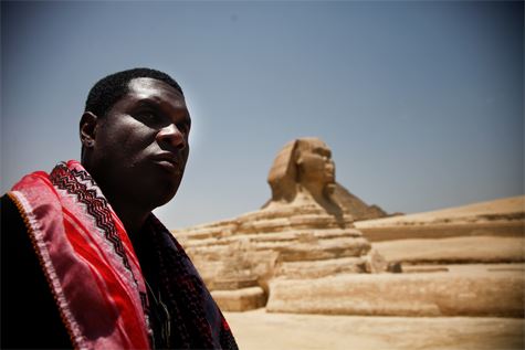 Jay Electronica standing in front of the Great Sphinx of Giza in Egypt, wearing traditional Middle Eastern clothing with a red keffiyeh