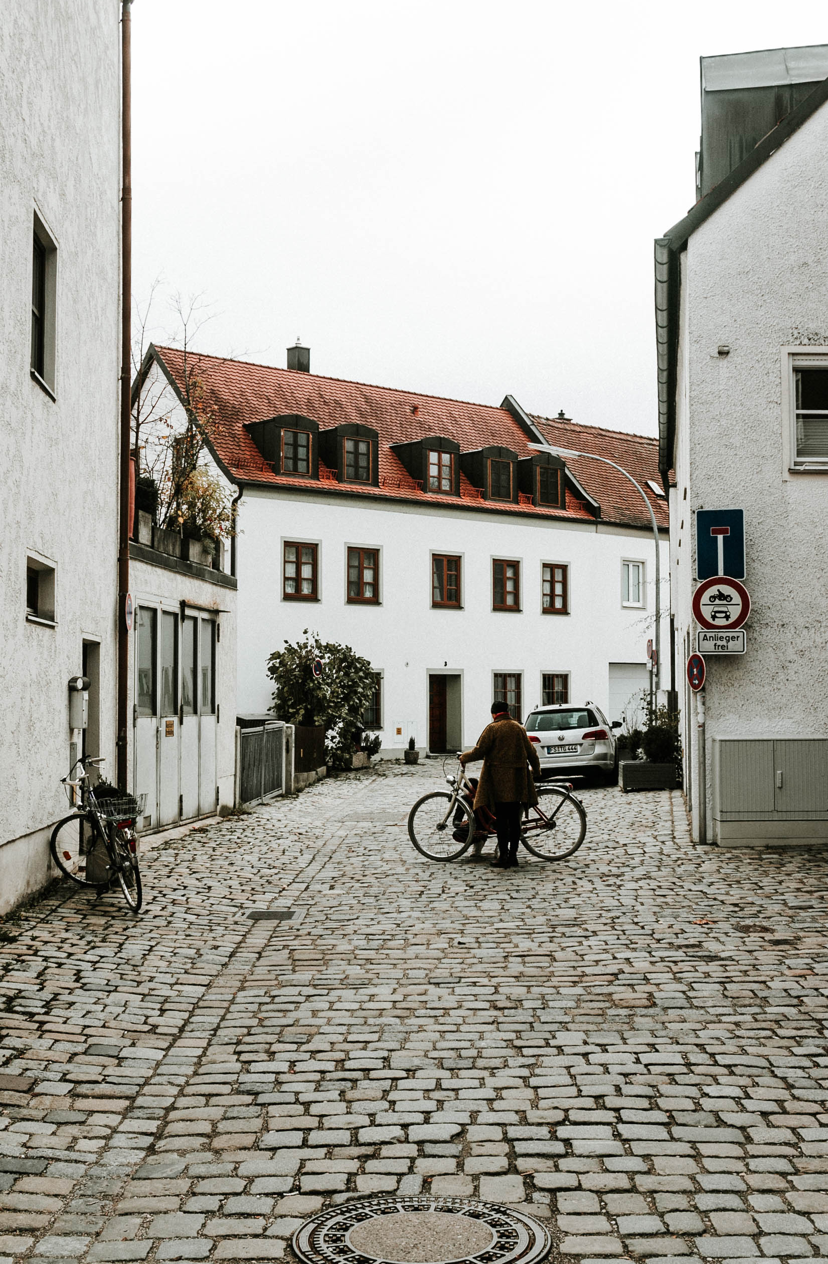 Just Passing Through - Person with bicycle on cobblestone street surrounded by traditional Bavarian architecture in Munich