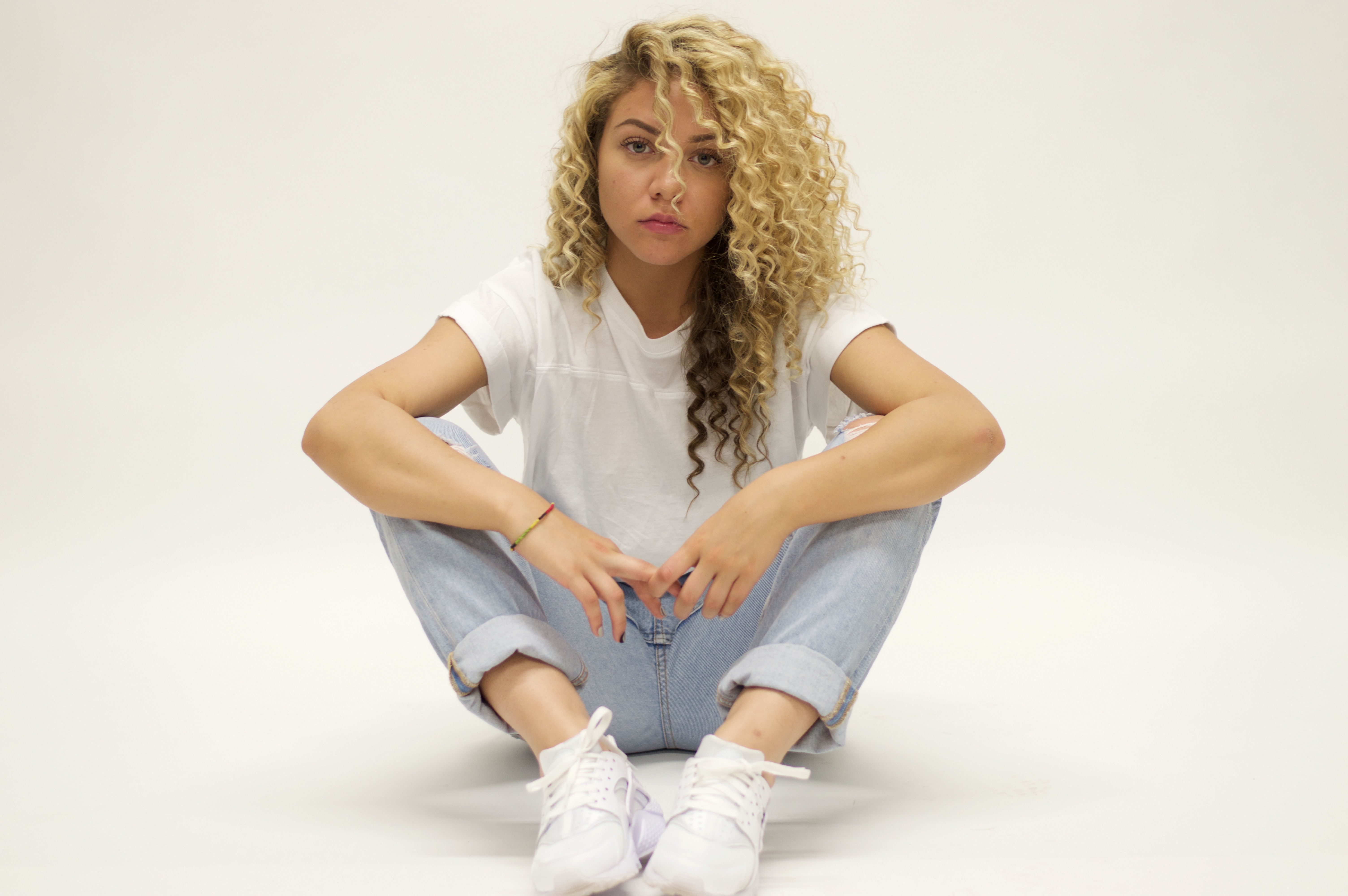 Let It Be Some Jaz Playing - Portrait of young woman with curly blonde hair sitting cross-legged in white t-shirt and jeans against white background
