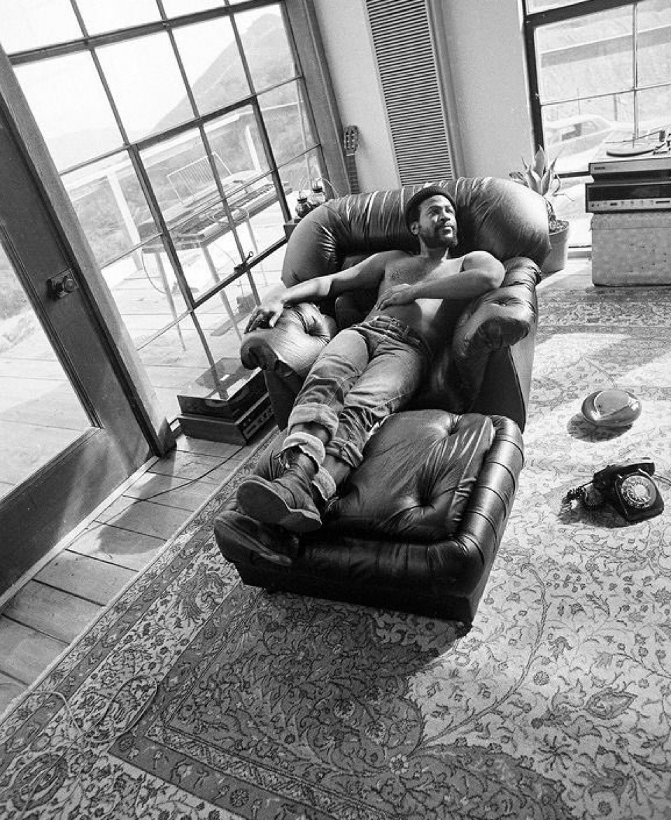 Black and white photograph of Marvin Gaye relaxing shirtless on a leather recliner in a sunlit room with large windows