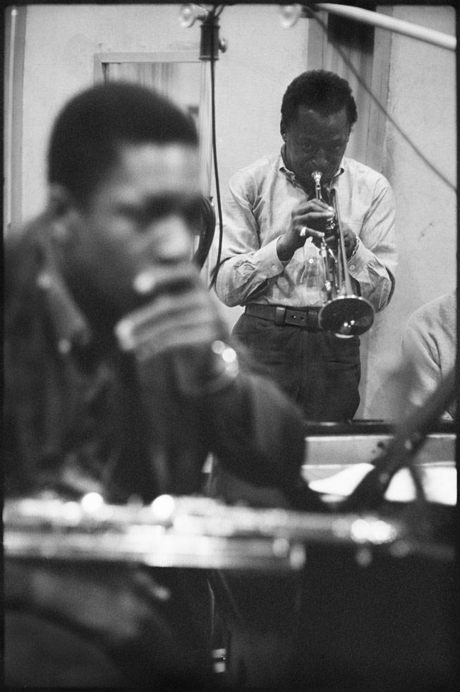 Black and white photograph from the Kind of Blue recording sessions showing Miles Davis in the background and John Coltrane in sharp focus playing trumpet in the studio