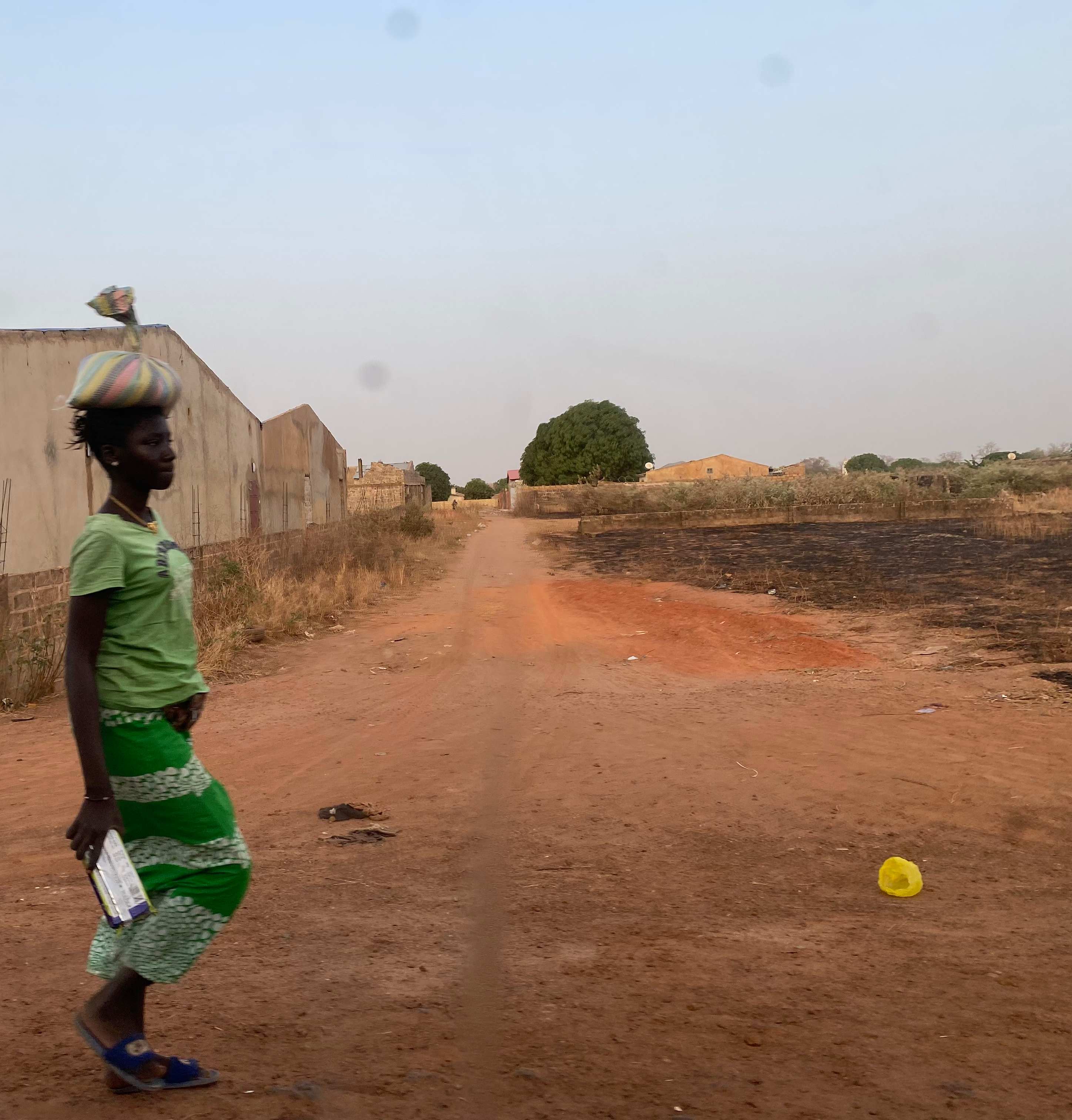 Senegalese Woman - Woman in traditional green dress carrying items on her head along a red dirt road in rural Senegal