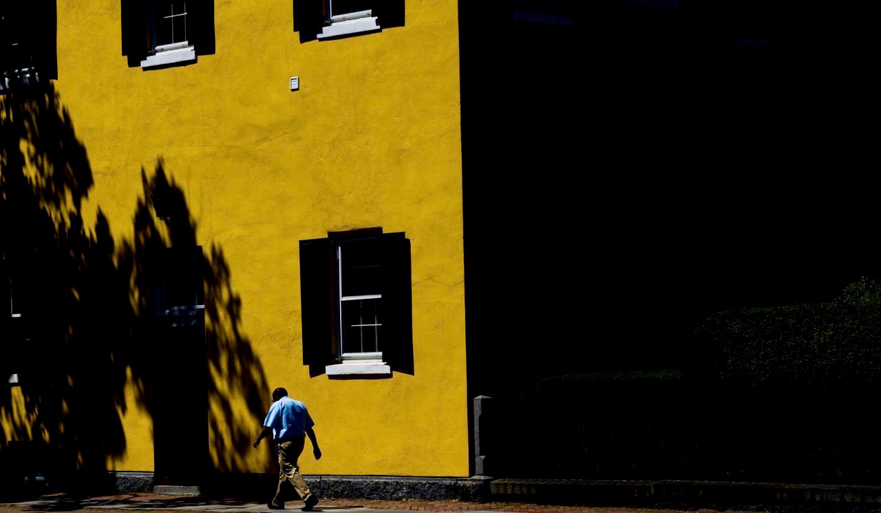 The Seeker - Person walking past a bright yellow building with dramatic shadows in Providence, RI