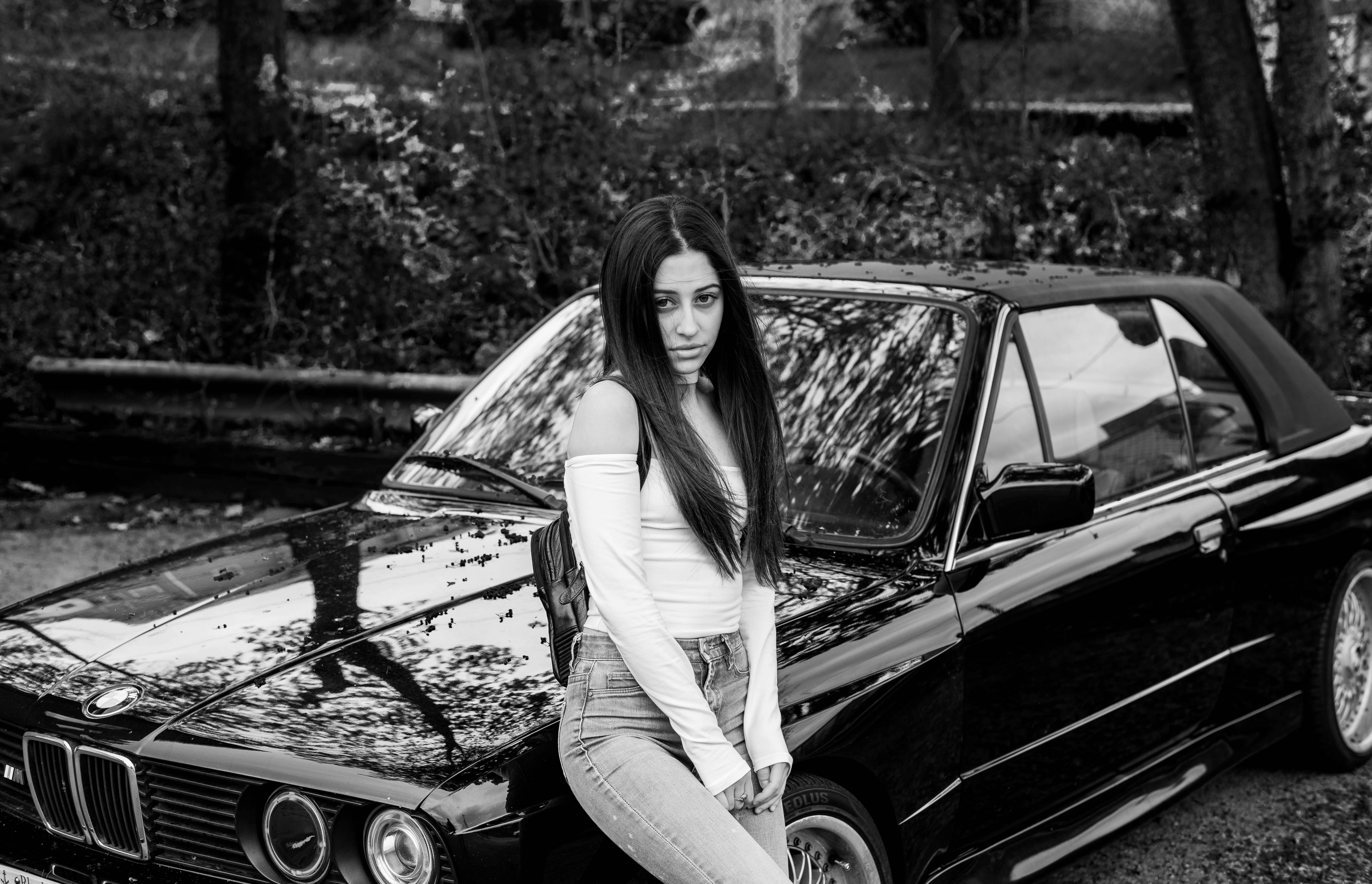 Whip Appeal - Black and white portrait of young woman with long dark hair posed against a classic BMW car
